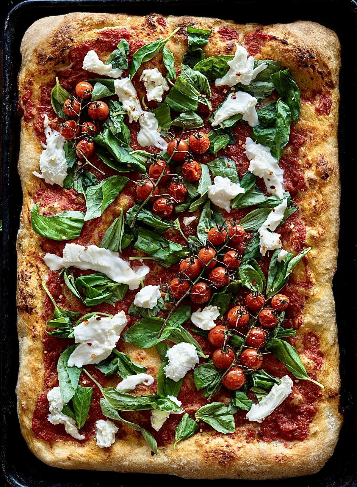 An XXL Roman-style tray pizza topped with tomato, basil, mozzarella and vine cherry tomatoes, baked on a black steel tray